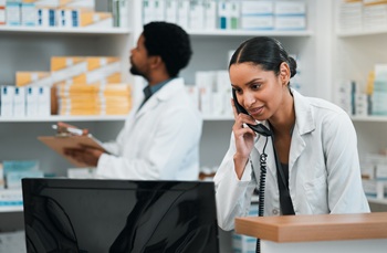 Two pharmacy employees - one on the phone and in the background one looking at medications on the shelves in the pharmacy.