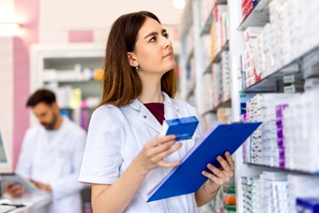 Pharmacy Clerk with clipboard in left hand and box of medication in right hand looking at shelf.
