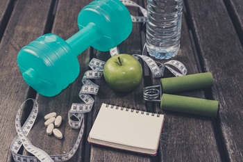 Image with a fitness theme - including a dumbbell, measuring tape, mineral water, fresh apple, hand grip, vitamin supplements & notepad or diary.
